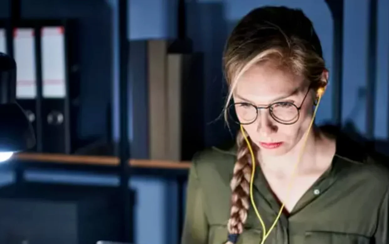 Konzentrierte Frau mit Brille und Kopfhörern, die nachts an beleuchtetem Schreibtisch arbeitet.