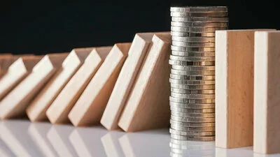 Liquidität symbolisiert durch aufgestapelte Münzen, die fallende Holzklötze in Reihe auf weißer Fläche stoppen.