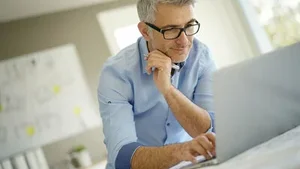 Factoring dargestellt durch älteren Mann mit Brille, der konzentriert an Schreibtisch über Laptop arbeitet und telefoniert.