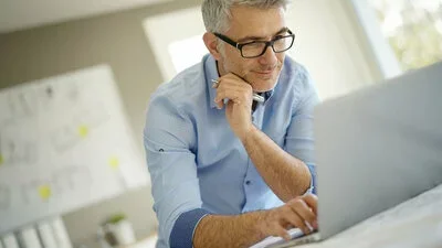 Factoring dargestellt durch älteren Mann mit Brille, der konzentriert an Schreibtisch über Laptop arbeitet und telefoniert.