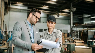 Leasing dargestellt durch Geschäftsmann mit Unterlagen im Gespräch mit Frau in Arbeitskleidung in großer Werkhalle.