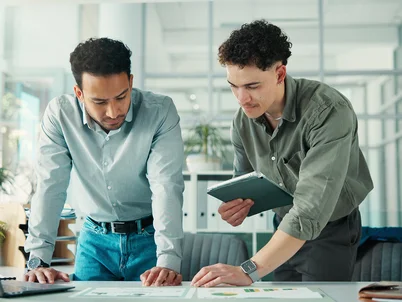 2 Männer schauen nachdenklich auf Skizzen auf dem Tisch neben einem Laptop