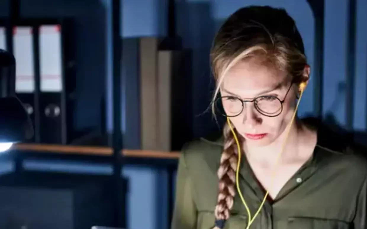 Konzentrierte Frau mit Brille und Kopfhörern, die nachts an beleuchtetem Schreibtisch arbeitet.