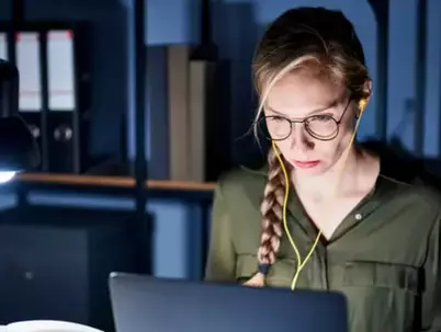Konzentrierte Frau mit Brille und Kopfhörern, die nachts an beleuchtetem Schreibtisch arbeitet.