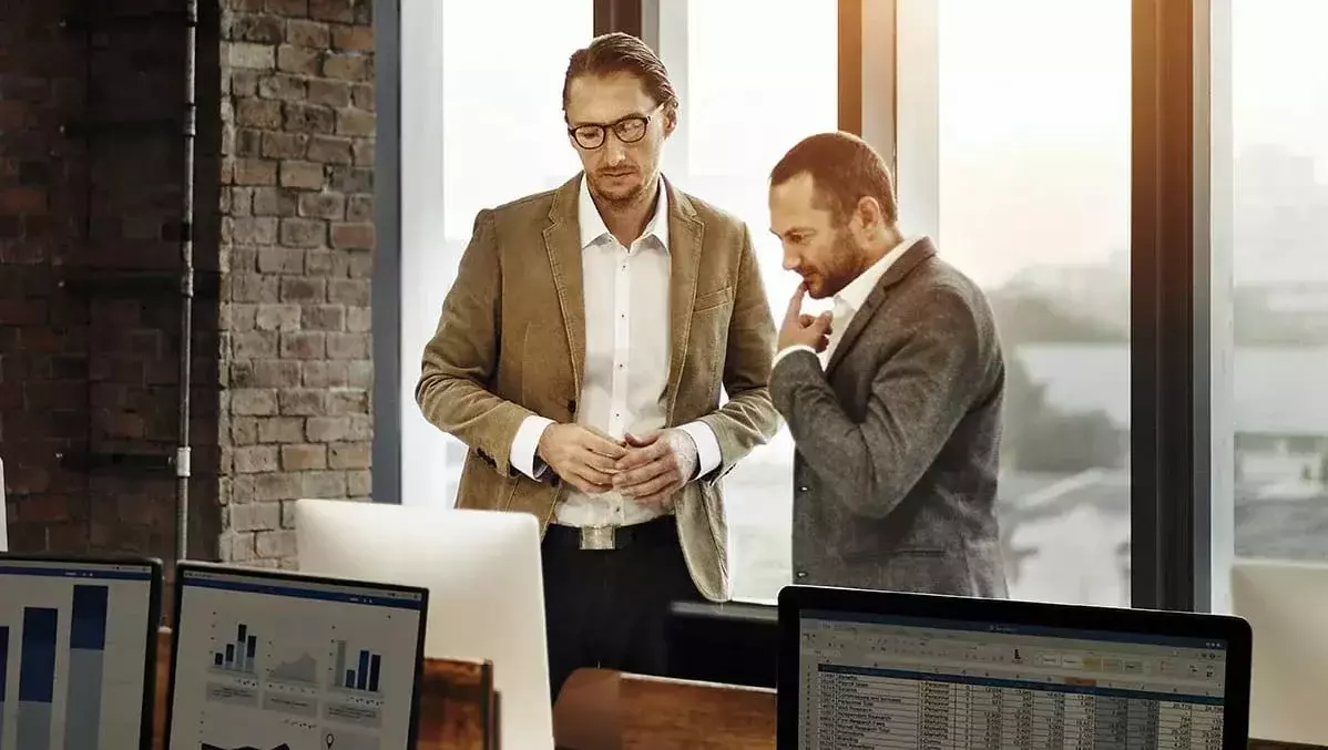 Zwei Business Männer sprechen im Office über Avalkredite, im Hintergrund Fensterfront, im Vordergrund Computer-Bildschirme-Desktops