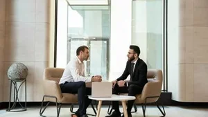 Finanzierung dargestellt durch zwei Männer im Business-Outfit beim Gespräch in heller, moderner Lobby mit Laptop.